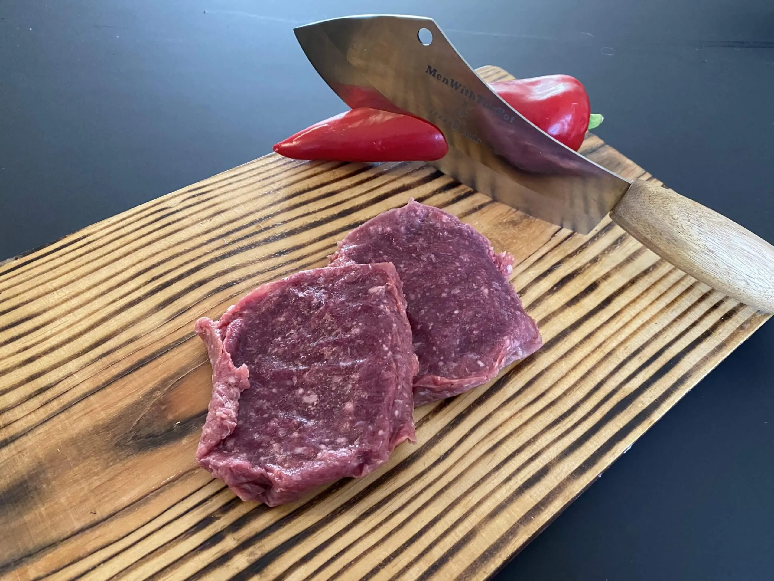 Twee steak tartaar op een houten snijplank, met een groot mes en een rode paprika op de achtergrond.